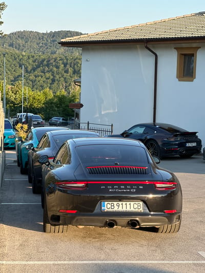 This is a portrait-oriented photograph taken outdoors, likely in Kerenite, Bulgaria. The scene features a line of parked cars, primarily Porsches, in a parking lot. The most prominent car in the foreground is a black Porsche 911 Carrera 4S, displaying the license plate "CB 9111 CB" and text indicating "PORSCHE OWNERS CLUB BULGARIA". To its left, a dark blue Porsche with the license plate "CB 7557 TK" is visible. Further back, other cars, including at least one turquoise vehicle, are parked in a row.

The setting includes a white building with a distinctive window and a tiled roof on the left side of the frame. Behind the cars and building, a lush green forested hillside dominates the background, suggesting a mountainous or hilly terrain. The sky is clear and bright, indicating a sunny day. The sunlight casts shadows on the pavement, suggesting it is either morning or late afternoon. The overall atmosphere appears to be relaxed, possibly during a car meet or a casual gathering of car enthusiasts. No people are visible in the image.