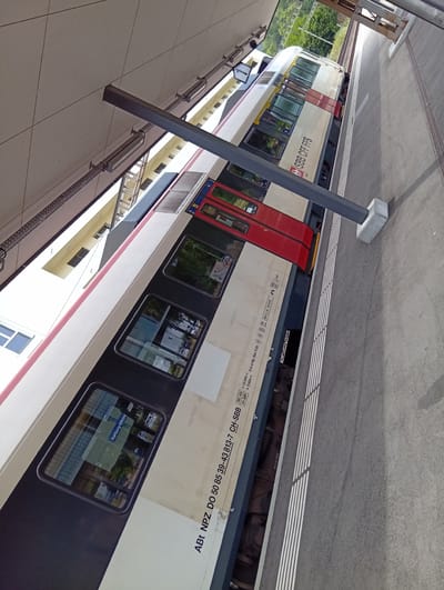 The image shows a close-up, angled view of a Swiss Federal Railways (SBB) train at a station. The train cars are predominantly white with a red stripe running along the side. The SBB CFF FFS logo is visible on one of the cars. Several windows are visible on the train, some reflecting the surrounding environment. One window displays "Aarburg-Oftringen" indicating the location. The train is positioned at a platform, with the track and platform edge clearly visible. A gray overhead structure supported by concrete pillars extends over the platform, providing shelter. The ground of the platform is paved and has a tactile warning strip. Lush green trees and foliage are visible in the background, suggesting a natural or semi-rural setting adjacent to the station. The lighting indicates it is daytime, likely sunny. No people are visible in the image. The overall scene depicts a typical railway station environment in Switzerland, specifically identified as being in Aarburg.
