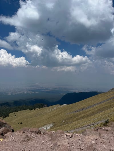 The image shows a landscape taken from a high altitude, possibly a mountain peak, overlooking what appears to be a valley or city in the distance. The right side of the image features a rocky, steep slope with grass. In the background, there is a valley with forest and a city, which based on the prompt, is the city of Guadaloupe, Mexico. The sky occupies the left side of the image, featuring clouds with patches of blue. The image is taken from a perspective with a visible horizon, suggesting a viewpoint from high above the ground.