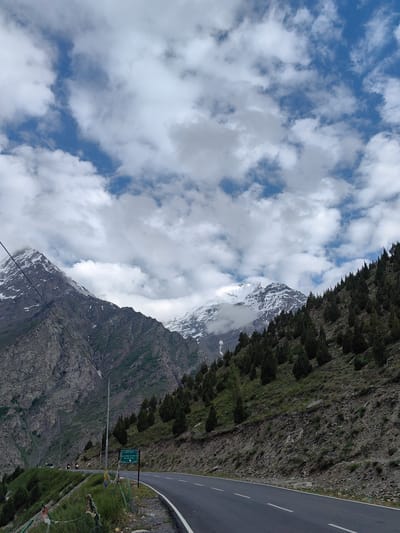 The image depicts a scenic mountain landscape with a winding road. The foreground features a paved asphalt road with white lane markings, curving to the right. To the left of the road, a grassy embankment is dotted with prayer flags. Further left, a steep, rocky mountainside rises dramatically, partially covered in greenery. A prominent signpost stands by the roadside, displaying text in a foreign script, likely Hindi, and a green background.
In the middle ground, the road continues into the distance, flanked by a lush green slope densely populated with coniferous trees. Beyond the trees, imposing snow-capped mountains dominate the background. The peaks are rugged and layered, with patches of snow and some clouds clinging to their upper reaches. The sky above is a mix of blue and white, with scattered, fluffy clouds creating a dynamic and somewhat overcast atmosphere.
A few distant vehicles, possibly motorcycles, are visible on the road, suggesting some level of activity or travel. The lighting indicates it is daytime, likely late morning or early afternoon, with the sun intermittently breaking through the clouds. The overall impression is one of a remote, natural, and somewhat majestic mountain environment, possibly in the Himalayas given the location context of Gemur, India. The scene evokes a sense of journey and the grandeur of nature.