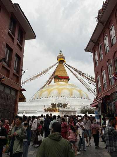 The image depicts the Boudhanath Stupa in Gokarneshwar, Nepal. The stupa, a large white dome with a golden tower adorned with eyes, is the central focus. Prayer flags are strung from the stupa's tower, extending towards the sky. The base of the stupa is surrounded by a crowd of people, many of whom are walking clockwise around the stupa. The people are wearing various styles of clothing. Buildings with brick facades flank the stupa on either side. The sky overhead is overcast.