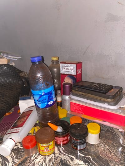 A cluttered shelf or tabletop displays various items. There's a stack of cellphones and cases, boxes of "White-Up" cream, and several jars of hair products with yellow, orange, and black lids. Two water bottles, one clear and one with a blue label, lie among the other objects. An orange-tipped item and other bottles and containers, some labeled "Demacol," add to the disarray. The background features a plain wall and a patterned surface.