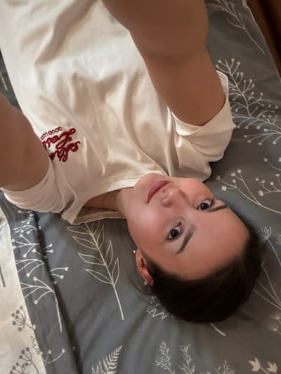 A female subject is observed lying supine on a bed in an indoor setting within the city of Bayronovka, Russia. She wears a white t-shirt with red embroidered text, including "good luck," on the left chest. Her dark hair is pulled back, and her dark eyes are directed upwards, engaging the camera. The bedding beneath her is grey with white botanical patterns featuring leaves and small flowering elements. Portions of her arms are visible, suggesting the image is a self-portrait.
