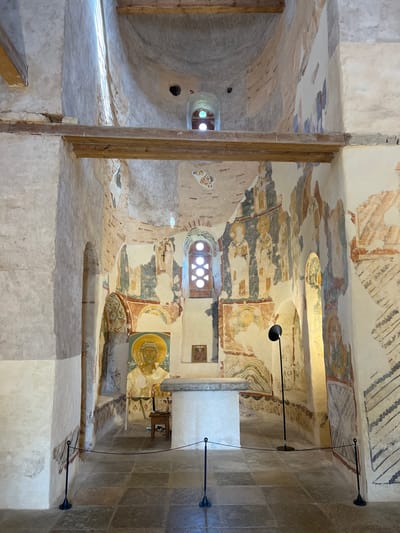 The image depicts the interior of the Church of the Transfiguration of the Savior, located in Spas-Nereditsy, Russia. The church's walls are adorned with frescoes, some of which are faded and damaged. In the center of the frame, an altar is visible, with a large fresco of a saint above it. A chair stands to the left of the altar, and a black floor lamp is positioned to the right. The floor is paved with square stone tiles. Wooden beams support the ceiling. Sunlight streams in from windows high up on the walls. The overall impression is one of ancient religious art and architecture.