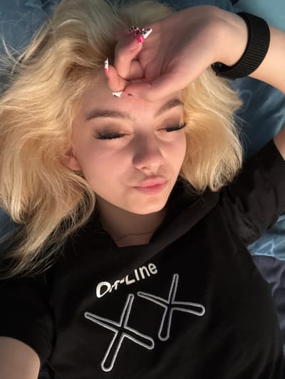 A young woman with blonde hair is lying down, covering her eyes with her hand. She is wearing a black t-shirt with white lettering and graphic design. She has long eyelashes and her lips are slightly parted. The background is a blue surface, possibly a bed or couch. The image does not provide any information about the city of Zhabokrat, Bulgaria.