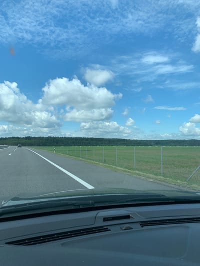 The image shows a view from inside a vehicle traveling on a paved highway. The highway has white lane markings and extends into the distance. To the right of the highway, there is a green field bordered by a fence. In the field, there are numerous round bales of hay. A dense forest lies beyond the field. The sky above is blue with scattered white clouds. The foreground shows the dashboard and windshield of the vehicle, including a windshield wiper. The image appears to be taken on a sunny day in a rural area. The location provided is Іўеўскі сельскі Савет, Belarus.