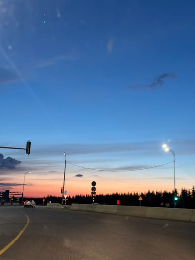 This image depicts a road scene at dusk, likely near Derzhavino, Russia. The sky displays a gradient of blues, transitioning to hues of orange and pink near the horizon. Silhouetted against the sky are numerous evergreen trees, forming a dense forest line. The road itself is paved, with a yellow lane marker visible. Traffic lights are present, with one showing a red signal and another a green signal. Streetlights illuminate the scene, and a few cars are visible on the road, one in motion. Concrete barriers line the side of the road.