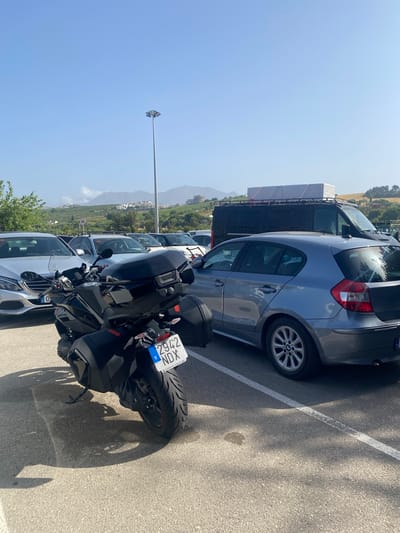A black motorcycle with saddlebags is parked in a lot. The motorcycle's license plate reads "2942 NDX". In the background, several cars are parked, including a silver Mercedes and a gray BMW hatchback. A black van with items strapped to its roof is also visible. The scene appears to be a parking area near a hilly, developed landscape with buildings scattered across the hillsides. The sky is clear and blue. This appears to be the parking lot of a commercial establishment in San Luis de Sabinillas, Spain.