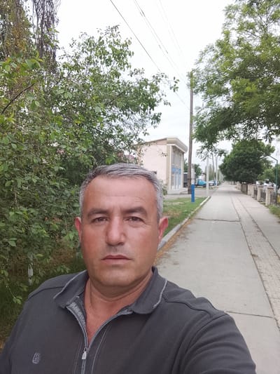 A selfie shot captures a middle-aged man in the foreground, looking directly at the camera with a neutral expression. He has short, greying dark hair, fair skin, and brown eyes. He is wearing a dark grey long-sleeved polo shirt with a half-zipper, and a small, unidentifiable emblem is visible on the left chest area.
The background shows an outdoor street scene, likely in Sabir Rakhimov, Uzbekistan, given the context. To the man's left and behind him are dense green bushes and trees, with some bare branches visible against the sky. To his right, a paved sidewalk stretches into the distance, lined on one side by more trees and low fences, and on the other by a grassy strip next to the road.
Further down the sidewalk, there's a light-colored building with some blue signage on its facade, though the text is unreadable. A utility pole with power lines runs parallel to the street, and a blue pole stands near the sidewalk. In the distance, other vehicles, including a light blue truck, are parked or driving. The sky is overcast, suggesting a daytime shot with soft, diffused lighting. The overall impression is a typical street view in a residential or semi-commercial area.