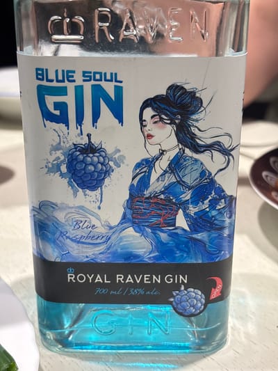 A close-up, eye-level shot shows the front label of a bottle of "ROYAL RAVEN GIN BLUE SOUL" positioned on a white, textured surface, possibly a table. The bottle is made of clear glass and filled with a vibrant blue liquid. The label features a stylized illustration of a woman with dark hair pulled back in a bun, wearing a blue kimono with white floral patterns and a red obi. To the left of the woman is a depiction of a blue raspberry surrounded by splashing liquid. The text on the label includes "BLUE SOUL GIN" in large, blue, drip-style lettering at the top, followed by "Blue Raspberry" in a script font. A black band across the middle of the label reads "ROYAL RAVEN GIN" with "700 ml / 38% alc." below it. To the right of this text is a small raspberry icon and a red arrow pointing to "OPEN." The brand name "RAVEN" is embossed in silver metal letters at the very top of the bottle. The background is blurred, with hints of a person's arm and shoulder visible to the right, suggesting an indoor setting. There is no specific information about the location in Падлабенскі сельскі Савет, Belarus in the image itself.