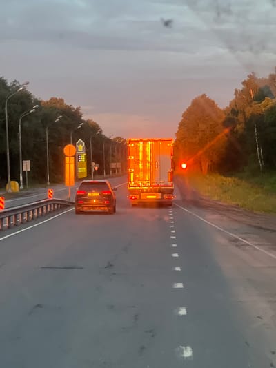 The image shows a road with two vehicles, a car and a truck, driving on it. The sun is setting, casting a warm orange glow on the scene. The truck's taillights are on, and the car's brake lights are also illuminated. The road is lined with trees on both sides, and there are streetlights visible along the side of the road. A gas station sign is visible on the left side of the image. The sky is partly cloudy, with the setting sun breaking through the clouds. The image appears to have been taken from a moving vehicle. There are no people visible in the image.