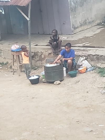 In the city of Alalubosa, Nigeria, a young person wearing a blue shirt is bending over a metal drum, seemingly washing clothes. Beside them, another individual sits on the ground. Nearby, an individual sits on a wooden stool holding a blue container. The ground is sandy and bare. In the background, there is a building with a grey wall and a corrugated metal roof. There are containers, including buckets and bags, strewn around.