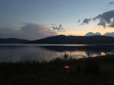 The image depicts a serene lakeside scene at dusk in Batak, Bulgaria. The sky transitions from a deep blue at the top to hues of pink, orange, and yellow near the horizon, with scattered clouds. Silhouetted against the colorful sky are rolling hills covered in trees. The calm lake reflects the sky and the distant hills.
In the foreground, the grassy shore is visible. A small campfire is burning, casting a warm glow. Adjacent to the campfire are two camping chairs, suggesting a leisurely outdoor activity. A fishing rod is planted in the ground near the water's edge. The overall atmosphere is one of tranquility and peacefulness, characteristic of a late evening by the water.