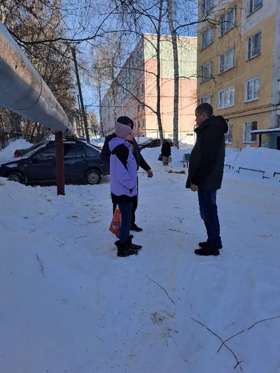 The image portrays an outdoor winter scene in Bolshaya Elhovka, Russia, under clear sky conditions with significant snow cover on the ground. Three individuals are central to the foreground. On the right, a male subject in a black hooded winter jacket, dark blue jeans, and black shoes stands facing left. Centered, a person in a light purple winter jacket, dark trousers, black boots, and a light-colored beanie holds a red bag in their left hand, facing right. A third individual, mostly obscured behind the central figure, wears a dark jacket and appears to be interacting with the other two. These three subjects are standing on a compacted snow path, suggesting an ongoing conversation.
In the mid-ground, a dark-colored sedan is parked on the left, partially covered in snow. A large, insulated utility pipe runs horizontally across the upper left quadrant of the frame. The background features two multi-story apartment buildings: one yellow with white-framed windows on the right, and another with alternating light green and pink sections further left. Bare trees, indicative of winter, are distributed throughout the scene. A solitary, distant figure in a light coat is visible near the yellow building on the right.