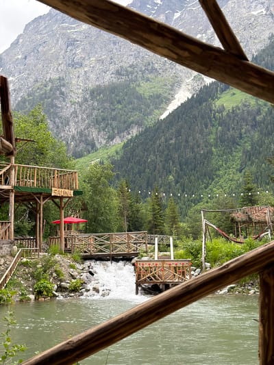 The image depicts a serene natural landscape with a body of water in the foreground. The water appears to be a river or a stream, with a visible current and some white water suggesting a small waterfall or rapids.
In the mid-ground, there are several wooden structures, including walkways, decks, and a building with a sign that reads "SAUNA." A red umbrella is visible on one of the decks. These structures are built over or near the water, and some are connected by wooden bridges. Lush green vegetation, including trees and bushes, surrounds the structures and the water.
In the background, a towering mountain range dominates the scene. The mountains are rocky and steep, with patches of green vegetation and some visible snow or ice at higher elevations. The sky is overcast, contributing to the soft, diffused light of the scene.
The image seems to capture a resort or a recreational area situated in a mountainous, natural environment, likely in or near Mazeri, Georgia, given the characteristic landscape and architecture. There are no people visible in the image. The overall impression is one of tranquility and natural beauty.