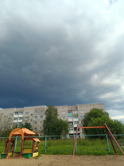 This media file captures an outdoor scene in Chusovoy, Russia, featuring a playground in the foreground and a large apartment building in the background. The playground includes a sandbox with a colorful play structure, predominantly orange and yellow, with the word "KVINTA" visible in text on its roof. A swing set with red metal supports is also present.

The scene is set against a backdrop of lush green grass and trees. The sky is dramatic, dominated by dark, heavy storm clouds that create a moody atmosphere. The lighting suggests it might be late afternoon or early evening, just before or during a storm. The apartment building is a multi-story, utilitarian structure, typical of Soviet-era architecture. There are no people visibly present in the image. The overall impression is one of an approaching storm over a residential area with a children's play area.