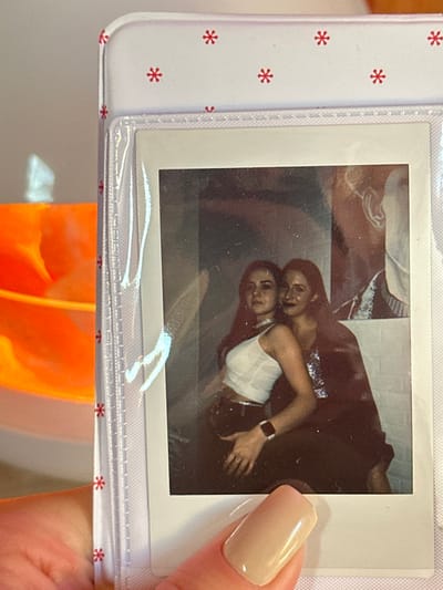 The scene depicts a human hand holding a white photo album page in 3-Y Uchastok, Russia. The album page features a repeating pattern of small red asterisk-like symbols and contains a single instant-print photograph secured within a clear plastic sleeve. The hand, with a short, nude-polished fingernail, grasps the bottom right corner of the album.
The instant photograph, a square-format print with a white border, displays two young adult females posing indoors. The female on the left has long dark hair, wears a white sleeveless top, a silver choker, and dark trousers. A dark smartwatch is visible on her left wrist, and her left hand rests on her hip. She looks directly at the camera with a neutral expression. The female on the right, also with long dark hair, is positioned slightly behind and to the right of the first subject. She wears a dark, possibly sequined top and has a slight smile or smirk. Her right arm is wrapped around the first subject's waist, with her hand resting on the left subject's left hip/buttock. The background of the photograph is dimly lit, revealing a dark, subtly textured wall behind the subjects. To the right, a large, partially visible graphic or artwork, appearing to be a stylized profile of a head with a prominent ear, is present in shades of brown and white.
Outside the photo album, to the left, a blurry, orange object with reflective highlights is visible. The wall behind the entire setup is light-colored and untextured.