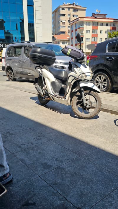 The image shows a street scene in Ataşehir, Turkey, on a sunny day. The foreground is dominated by a parked white scooter with a black storage box on the back and a blue bag with reflective stripes on the handlebars. The scooter is parked next to a gray van. In the background, there are multi-story buildings with apartments and shops, one of which has a blue glass facade. The street is paved with gray tiles. The sunlight casts long shadows across the pavement, indicating it is likely late afternoon. The scene is calm and depicts everyday urban life. No people are clearly visible in the image, but a foot wearing a sandal and casual trousers is visible in the bottom right corner. No text is legible in the image.