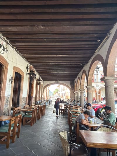 An outdoor corridor displays a series of arches along the top. The left side features a dark, wooden structure with vertical planks. Tables and chairs line the right side of the corridor, where a group of people are seated near the top of the image. A person walks along the corridor further down, while more seating arrangements are visible in the distance leading to the open end with daylight.