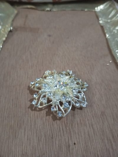 A silver-toned metallic brooch, intricately designed in a multi-layered floral pattern, rests centered on a light brown wooden surface with visible vertical grain. The brooch is adorned with numerous clear, faceted rhinestones of varying sizes, with a prominent central stone displaying iridescent reflections. A distinct shadow is cast to the immediate right of the object. The background features blurred areas of shiny, yellowish-gold material along the top and right edges of the frame. No individuals are present, and no actions or interactions are depicted. The scene is static, located in Chandragiri, Nepal.