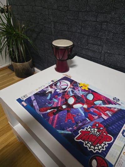 This image displays an indoor scene located in Zweibrücken, Germany. The central foreground features a white, two-tiered rectangular table or shelf unit. On its top surface, a partially assembled jigsaw puzzle depicting characters from the Spider-Man universe, specifically Spider-Gwen, Miles Morales (Spider-Man), and another Spider-Man variant, is laid out. The puzzle's artwork shows the characters against a dark urban backdrop with vibrant neon lights. Visible text on the puzzle includes "SUPER HEROES" and "49" (likely indicating piece count), along with a red logo featuring a spider and "TEAM SPIDEY" style lettering. To the left of the puzzle, a small, dark red djembe drum rests, equipped with a light-colored drumhead secured by dark lacing, and a black cord or strap hanging from its side. The background wall is covered with dark grey, textured wallpaper designed to mimic stacked stone or brickwork. To the far left, a potted plant with multiple slender stems and green leaves is situated in a dark, round pot. A light-colored wooden floor is visible beneath the furniture in the lower left portion of the frame. No individuals are present, and no active interactions are depicted.