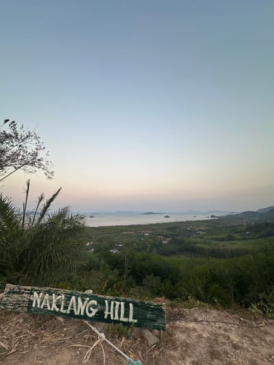 The image captures a panoramic view from Naklang Hill in Ko Lanta Yai, Thailand, at what appears to be twilight. In the immediate foreground, a weathered, horizontally oriented green wooden sign with raised white letters spelling "NAKLANG HILL" rests on reddish-brown, dry soil. A white rope, possibly knotted or coiled, lies on the ground to the left of the sign. To the left and slightly behind the sign, dark, sparse tree branches with small leaves and several prominent green palm fronds partially frame the scene.

Beyond the foreground, the landscape descends into a dense expanse of lush green vegetation, indicative of a tropical forest or plantations, covering rolling terrain. Scattered structures with light-colored rooftops are visible amidst the greenery in the midground. Further into the distance, a calm body of water, likely the sea, stretches horizontally across the frame, punctuated by several small, green-covered islands. Distant landmasses or additional islands are faintly visible along the horizon line. The sky, occupying the upper two-thirds of the image, displays a subtle gradient, transitioning from a light blue at the zenith to a pale pink and orange hue near the horizon. No individuals or active subjects are present.