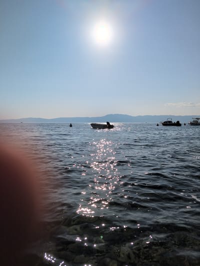 The image depicts a sunny day scene at sea, likely on the coast of Kraljevica, Croatia. The foreground features choppy, dark blue water with sunlight reflecting off its surface, creating a sparkling effect. Partially submerged rocks are visible beneath the water's surface in the lower right corner. In the mid-ground, several boats are anchored or moored on the water. One is a small, white motorboat with a dark hull, positioned centrally. To the right, two larger motorboats are visible, one with a canopy. A black buoy is also present in the mid-ground, to the left of the central motorboat. The background shows a hazy, mountainous coastline under a clear, pale blue sky. The sun is high and bright, casting a glare in the upper portion of the image. A blurred, reddish-brown object, possibly a hand or part of a person, is visible in the extreme lower left corner, suggesting the photo was taken from a close vantage point near the water's edge. The overall impression is one of a calm, bright day at sea, with recreational boats present.