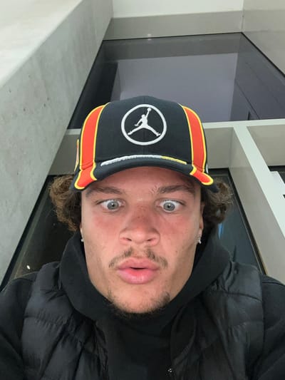 A selfie of a man is taken indoors with an unknown location in Ivry-sur-Seine, France. The man is fair-skinned with light-colored eyes and short brown curly hair. He is wearing a black baseball cap with the Jumpman logo and a red and orange stripe around the edge. He is also wearing a black hoodie and a black puffy jacket. The background is blurry but appears to be a modern interior with grey and white walls and a window or glass panel.
