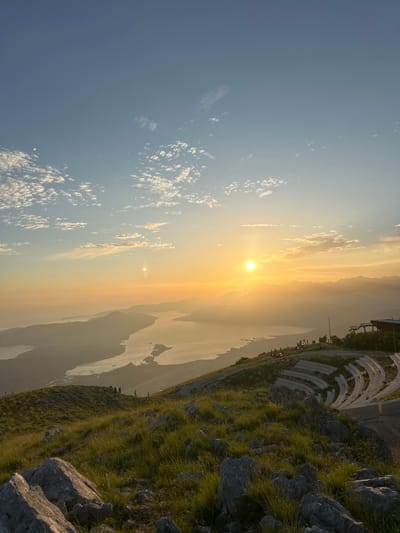 An aerial, wide-angle view captures a sunset over the Bay of Kotor, from a vantage point in Kavač, Montenegro. The sun, a bright orb, hangs low in the sky, casting a warm golden-orange hue across the scene and illuminating scattered clouds. The bay, with its tranquil waters, is nestled between rolling hills and distant mountains, all softened by a gentle haze. On the right side of the image, a tiered outdoor amphitheater is visible, carved into the hillside, with stone seating arranged in curved rows. A pathway leads up to the amphitheater, and a ski lift structure is present near the top of the hill. The foreground is dominated by the rugged texture of a grassy hillside, dotted with large rocks and illuminated by the fading sunlight. Several individuals are scattered across the landscape, some walking along the hillside and others seated in the amphitheater, enjoying the panoramic view. The overall impression is one of serene natural beauty and a peaceful gathering space.