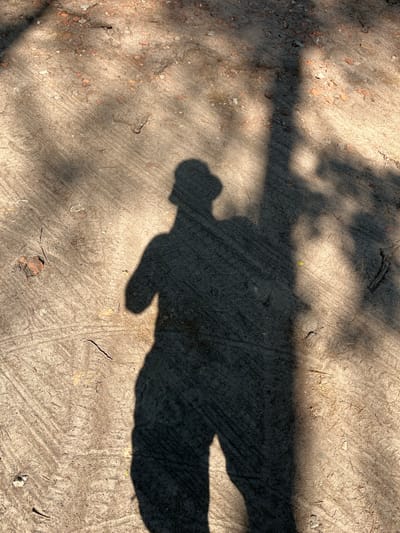 This image captures the shadow of a person cast on a sandy, textured surface. The shadow is elongated, suggesting it's midday or late afternoon with the sun low in the sky. Sunlight filters through overhead foliage, creating dappled light and shadow patterns on the ground, hinting at an outdoor setting, possibly a park or a natural area. The ground appears dry and gritty, with small pebbles and dried plant debris scattered across its surface. The shadow itself is sharp and well-defined, showing the silhouette of a person with a rounded head and a body extending to the right. A faint, lighter shadow, possibly from an arm or an object held by the person, extends from the main shadow. The overall impression is one of a moment captured in time, with the interplay of light and shadow dominating the scene. The location context of Zalesie Górne, Poland, provides a geographical setting, but offers no specific visual cues within the image itself. There is no visible text in the image.