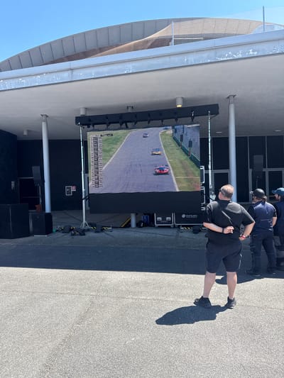 Here is a detailed description of the image:

The image appears to be taken outdoors on a sunny day in Campagnano di Roma, Italy. A large LED screen displays what looks like a car race. People are standing and watching the screen. The screen is set up against a building with a curved roof structure overhead. The ground is paved with asphalt. Cables and equipment are connected to the LED screen. There is a black stage in front of the screen.