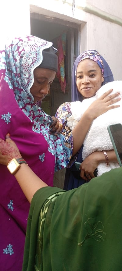 The image depicts three individuals in an outdoor or semi-outdoor setting in Dan Maliki, Nigeria. In the center-left, an adult woman with dark skin wears a dark cap under a magenta head covering featuring light blue floral embroidery; she looks downwards, her right hand, adorned with henna, is positioned near her chest. A gold-framed watch with a dark strap is visible on her left wrist. To her right, a younger woman with lighter skin, wearing a blue floral head covering and a nose piercing, smiles slightly towards the viewer while holding a white, fluffy object, likely a swaddled infant, with both hands. Her hands also display henna designs, and a rose gold bracelet is present on her left wrist. Partially visible in the lower foreground, an arm dressed in a dark green embroidered garment holds a black smartphone, indicating the perspective of the photographer. The background consists of a light-colored wall and a dark doorway, within which patterned fabric is visible.