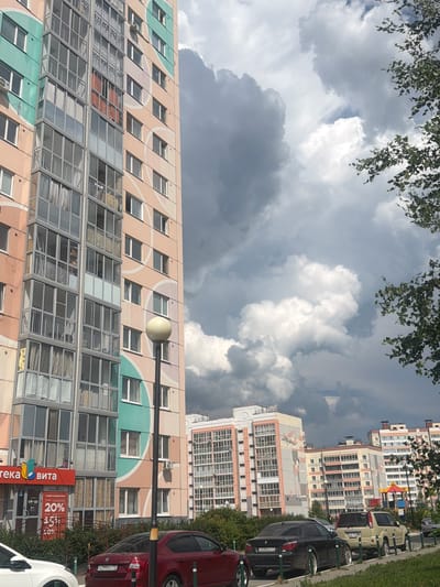 This is a vertically oriented, eye-level, outdoor medium shot of a residential building with a complex facade featuring balconies and windows. The building is tall and appears to be in an urban or suburban setting. The facade is adorned with pastel-colored geometric patterns and large circular motifs in shades of pink and teal. Several stories of the building are visible, with some windows open and others closed. The lower levels of the building show more modern additions, including what appear to be enclosed, glass-fronted balconies or sunrooms.

In the foreground, on the right side of the image, are several parked cars, including a red car and a black car. The black car has a license plate that reads "A038EO 77 RUS". A white car is partially visible at the very bottom right of the frame. There is a street or sidewalk running alongside the building and cars, bordered by a grassy area and some trees.

Above the building, the sky is filled with dramatic, dark gray storm clouds interspersed with patches of bright white, fluffy clouds. Sunlight is breaking through in some areas, indicating it might be a partly cloudy day with potential for rain. A few green branches with leaves hang from the top of the frame, suggesting trees are present.

Visible text includes "20%" and "08-22" on a red sign attached to the building, possibly indicating a sale or opening hours for a store. There is also text on other signs, including "ВИТА", which appears to be a brand name, and "Аптека" (Apotheka, meaning Pharmacy).

The overall impression is of a residential area on a day with mixed weather conditions, transitioning from sunny to potentially stormy. The building's colorful facade adds a lively element to the scene.