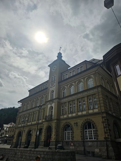 This media file captures an upward-angled view of an old building with a prominent clock tower. The building, with its pale yellow facade and arched windows, appears to be situated in a European town. The clock on the tower shows the time as approximately 3:50. The sky is overcast with dramatic, textured clouds, and the sun is visible as a bright, hazy orb, suggesting either early morning or late afternoon. The scene is one of a historical urban environment, possibly in St. Moritz, Switzerland, given the context. There is a street with cobblestones in the foreground and the architecture suggests a place with a rich history. The presence of streetlights and what appears to be a traffic sign with German text, reading "Vorsicht Rutschig," which translates to "Caution Slippery," further hints at a European location. A few indistinct figures are visible on the street, and a person with red clothing is seen in an arched doorway, but their activities are not clearly discernible. The overall mood is somewhat dramatic due to the sky and the angled perspective.