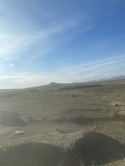 The image captures a vast, open landscape under a clear, bright blue sky with wispy white clouds stretching across. The terrain consists mainly of dry, light brown earth and extensive areas covered in small stones or gravel, interspersed with sparse patches of green vegetation.
In the midground, several low-lying structures, possibly small buildings, sheds, or informal dwellings, are visible against a backdrop of distant, hazy hills or mountains on the horizon. The ground in the foreground and midground is traversed by what appear to be unpaved tracks or pathways. Throughout the scene, thin poles and wires suggest fences or boundary markers. A distinct feature is a fence in the foreground with a piece of white and red fabric, possibly a flag, attached to it. Scattered debris can be seen on the ground in various places. A large, dark shadow falls across the right side of the foreground, suggesting an object or structure just out of frame.
The scene is bathed in bright daylight, with clear illumination and shadows, indicating a sunny day. No people are visible, and no specific activities are taking place, contributing to a sense of stillness and openness. The environment appears rugged and rural, aligning with the provided location context of Jbilate, Morocco. There is no discernible text within the image.