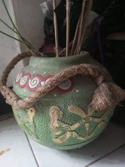 An aged green vase with a brown rope handle sits on a white tiled surface. The vase features red and white swirl designs near the top, with carved golden figures adorning the lower portion. Inside the vase are green sprigs and thin wooden rods, possibly decorative. The rope handle, adorned with a tassel on one side and tied with a bamboo piece on the other, adds to the vase's rustic aesthetic.