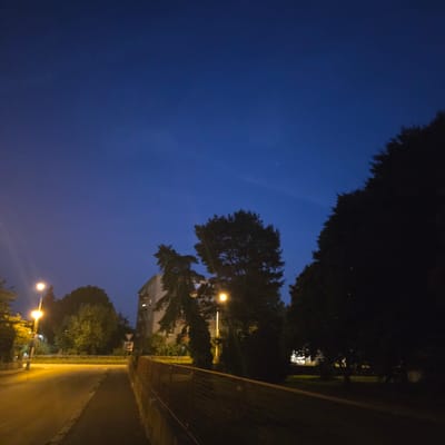 The image depicts a street scene at dusk or night in Piešťany, Slovakia. A street lamp illuminates a paved road on the left, which curves into the foreground. To the right of the road, a low fence with a wire mesh runs parallel to a sidewalk or pathway. In the mid-ground, trees are silhouetted against a dark blue sky. Behind the trees, a multi-story building with visible windows is partially illuminated. Another street lamp is visible between the trees and the building. The sky is clear with a faint wispy cloud. There are no people visible in the image. The overall atmosphere is quiet and urban.