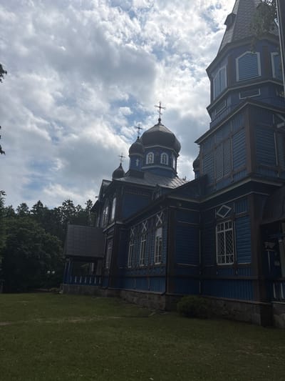 The image shows an exterior view of the Orthodox Church of the Nativity of the Blessed Virgin Mary in Puchły, Poland. The church is made of wood and is painted blue with dark brown trim. It has multiple onion domes topped with crosses. The church is situated on a grassy area with trees in the background. The sky is cloudy. The perspective is from the side of the church, looking up towards the domes and spire. The building's architecture features decorative window frames and horizontal wooden siding. The base of the church appears to be made of stone. There is a porch or entryway visible on the left side of the building.