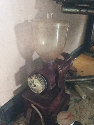 An electric coffee grinder, dark red/maroon in color, is centrally positioned against a light-colored, textured wall in Dogsenang Gorphoog, Bhutan. The grinder features a clear, conical plastic hopper on top, which appears dusty and has two small vertical protrusions. Below the hopper, a silver-colored, circular grind adjustment dial with a textured edge and "MEDIUM COARSE FINE" markings is visible on the front of the grinder's body. The base of the grinder rests on a dark, possibly concrete or tiled, surface that shows some dust and debris.
To the right and behind the grinder, dark, horizontally oriented cylindrical objects (possibly pipes or bars) are visible, along with a piece of light brown material resembling cardboard. On the dark surface in the lower right foreground, a small, light-colored cylindrical object lies horizontally, and a circular object with a red indicator mark is partially visible. A white cable extends from the lower left side of the grinder's base. Strong shadows are cast on the light-colored wall behind the grinder, indicating a direct light source. No people or active interactions are depicted in the scene. The overall environment appears functional and somewhat unkempt.