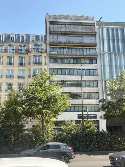 The image captures a street view in Neuilly-sur-Seine, France, on a clear, sunny day. The foreground features a street with two cars, one dark gray SUV on the left and a portion of a silver car on the right. Beyond the street, a row of buildings dominates the scene. The building on the left is a light-colored apartment building with traditional Parisian architecture, featuring mansard roofs, dormer windows, and balconies with black railings. Adjacent to it, to the right, is a modern, taller building with a concrete facade and numerous horizontal windows, some with closed blinds. Further to the right is a building with a striking facade composed of vertical, translucent panels. Lush green trees with dense foliage partially obscure the lower portions of the buildings. A streetlamp stands prominently in front of the concrete-faced building. The upper floors of the buildings show balconies and rooftop structures, one with greenery and what appear to be flowering plants. Signage on the ground floor of the concrete building indicates "CENTRE MEDICAL & DENTAIRE" and "HTALILOGIE," suggesting it houses a medical and dental center.