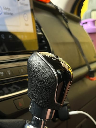 The image shows a close-up view of a car's interior, focusing on the gear shifter and a portion of the dashboard with a smartphone. The gear shifter is black with a textured grip and a metallic trim, featuring the letters "R", "N", and "D" visible on its top surface, indicating it's for an automatic transmission. A white charging cable is plugged into a black adapter located behind the gear shifter, suggesting the smartphone is being charged. The smartphone displays a screen with some text in what appears to be German, including "Telefon" and "Verbinden," which translate to "Telephone" and "Connect," respectively. The overall setting is inside a car, with the background showing other car interior elements and possibly a blurred view of the outside. The lighting suggests it could be daytime, though the exact time is unclear. There are no people visible in the image. The location is identified as Neuss, Germany, but there are no specific landmarks visible to confirm this.