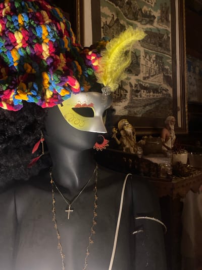 A close-up photograph features a black mannequin adorned with various accessories and decorations. A masquerade mask with a yellow feather rests atop a colorful, textured garment resembling yarn. A gold-colored necklace with a cross pendant and a second necklace decorated with star shapes are draped around the mannequin's neck. The mannequin also wears jewelry, including what appears to be a beaded bracelet. In the background, a framed image and other decorative objects are partially visible. The location is Baan Don Sai, Thailand.