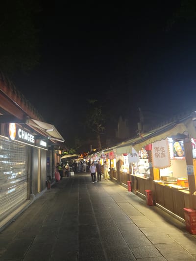 This is a nighttime, eye-level, full-shot photograph of a bustling pedestrian street lined with food stalls and shops in Wuhou District, China. The street is paved with dark, rectangular stones, and illuminated by the warm glow of lights from the stalls and street lamps. The sky above is dark, with a hint of tree foliage visible in the upper left corner.
On the left side of the street, a store with a prominent "CHAGEE" sign is visible. Its entrance is covered by a metallic roll-down gate. Further down the street, several figures are present, including a security guard wearing a reflective vest, and a few pedestrians browsing at the stalls.
The right side of the street is dominated by a row of food stalls. These stalls have wooden counters and are adorned with various signs, some featuring Chinese characters and images of food items. Red lanterns hang from some of the stall awnings. People are seen milling around the stalls, presumably purchasing or consuming food. The stalls are brightly lit, showcasing food displays and cooking equipment. Red cylindrical trash cans are placed along the sidewalk in front of some of the stalls. The overall atmosphere is lively and vibrant, characteristic of a busy night market.