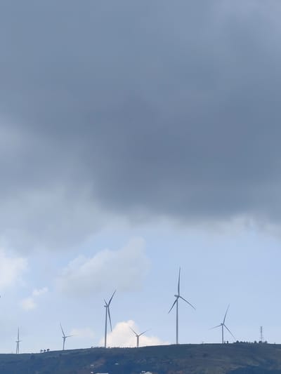 The image depicts a wind farm located in Đa Thọ, Vietnam. Several wind turbines stand on a grassy hillside under a sky filled with thick, dark clouds. The turbines are of varying heights and distances from the viewer. Structures, possibly houses, can be seen at the base of the hill. An electrical pylon is visible at the far right of the hillside. The overall impression is of a cloudy, possibly stormy, day.