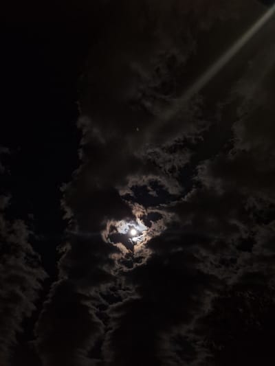 The media file presents a dramatic night sky filled with dark, textured clouds over Dolni Chiflik, Bulgaria. A prominent, intense light source, likely the moon, is partially visible through a break in the clouds in the upper-middle right portion of the image. This light casts a bright, soft glow, illuminating the edges of the surrounding dark cloud formations and creating a subtle halo effect.
A distinct, straight beam of bright light diagonally traverses the sky from the upper-left corner downwards, cutting through the darkness and clouds. Its origin is outside the frame. The clouds vary in density, with some appearing as heavy, opaque masses and others allowing a glimpse of the dark sky behind them.
The overall color palette is dominated by deep blacks and various shades of dark gray for the sky and clouds, sharply contrasted by the bright white and subtle yellowish hues emanating from the celestial light source. Small, circular green lens flares are also visible in the image, one near the prominent light beam in the upper left, and another smaller one closer to the moon, indicating a night-time photograph with a direct light source.
The scene captures a serene yet mysterious atmosphere, characteristic of a cloudy night. There are no people, man-made objects within the clouds, or visible text in the image. The time of day is clearly night.
