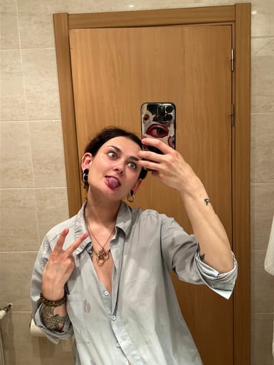 A woman is taking a selfie in a bathroom. She is holding her phone in her right hand, which has a phone case with eyes on it. The woman has dark hair, green eyes, a nose piercing, and stretched earlobes with hoop earrings. She is wearing a light gray button-up shirt and several necklaces. Her tongue is sticking out, and she is making a peace sign with her left hand, which has tattoos and a beaded bracelet. The bathroom has light brown wooden doors and beige tile walls. There is no evidence to suggest the location is Puçol, Spain.