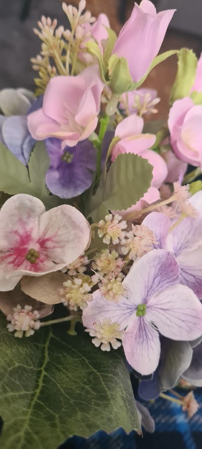 A close-up photograph captures a vibrant bouquet or arrangement of artificial flowers, showcasing a variety of floral types and colors. The arrangement primarily features shades of pink, lavender, light purple, and white, complemented by green leaves and stems. Prominent elements include soft pink, closed rosebuds with green sepals.

Among the open blossoms are fabric flowers with several distinct petals, some appearing light lavender with purple veining and a green center, resembling hydrangeas. Other open flowers are pale pink or white with bright pink centers and green stamens. Scattered throughout are clusters of small, textured, off-white to pale yellow filler flowers, some with tiny reddish specks, contributing to the arrangement's depth. Green leaves with visible venation and serrated edges are interspersed, adding a natural touch.

The visible textures confirm the flowers are artificial, likely made from fabric. The background is softly blurred, revealing hints of a dark blue, possibly patterned or plaid, fabric on the right side, suggesting an indoor setting. No human activity or visible text is present in the image. The scene is set in Beringen, Belgium, although no specific visual cues within the image identify this location.