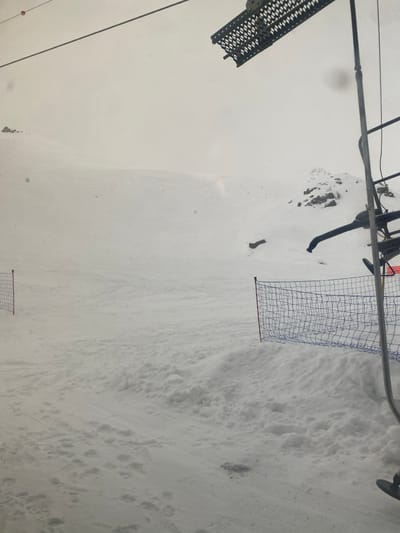 The scene depicts a snow-covered mountain environment in La Salle-les-Alpes, France, under an overcast, light grey sky. In the immediate foreground on the right, components of a ski lift are visible, including a dark metal support pole, a portion of a black chairlift seat, and an overhead dark grey grated platform structure. The midground is dominated by a broad expanse of disturbed snow, displaying various ski tracks and uneven terrain. Two distinct segments of safety netting are present: one on the left and one on the right. Both nets are blue with a red border, supported by vertical metal poles, and define sections of the snow-covered slope. The background consists of ascending snow-covered slopes, with several dark rocky outcrops visible through the snow on the right side, fading into the uniform light grey of the overcast sky. No human subjects or active events are present.
