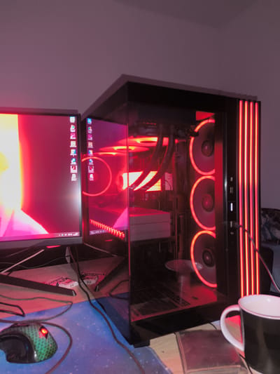 This media file depicts a personal computer setup in a dimly lit room, suggesting evening or nighttime. The scene is dominated by vibrant red lighting emanating from the computer components and monitor screen, creating an immersive atmosphere for gaming or general use.
The central subject is a modern gaming PC featuring a transparent side panel that reveals its internal hardware. Multiple fans with glowing red rings are visible on the side, alongside vertical red light strips on the front panel. Internal liquid cooling tubes and other components are also bathed in red light.
To the left, a large monitor displays a dynamic red and orange background, reflecting the PC's red glow. A column of application icons, including browsers, communication tools, and media players, is visible on the screen's left edge. The taskbar shows the time "20:50".
On the desk, a wired gaming mouse with a green, scaly texture and a glowing red element is positioned on a blue and purple cosmic-themed mouse pad. A black mug with a white handle and rim is situated to the right. Underneath the monitor, two white boxes distinctly labeled "Energizer" are visible. Multiple black and brown cables are present across the wooden desk surface. A small, dark book or box with a textured cover is also on the desk. The background consists of a plain wall, with a hint of patterned fabric. This setup is located in Havixbeck, Germany.