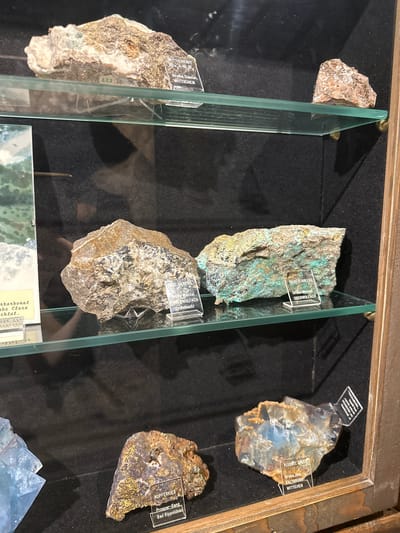 The image displays a collection of mineral specimens in a museum display case, likely located in Schonach im Schwarzwald, Germany, as indicated by labels on some of the specimens. The display case has glass shelves with several rocks and minerals.
On the top shelf, a rock labeled "WITTICHENIT Cu3 BiS3" is visible, along with another unlabeled rock on the right.
The middle shelf holds two larger specimens. The left specimen is a roughly textured rock with lighter and darker brown and gray tones. The right specimen is a large, irregular chunk of rock with prominent green and turquoise mineral deposits. Both have small, clear plastic labels. The left label is partially obscured but seems to read "GEIL SILBER" and "Grube Wenzel OBERWOLFACH (Frohnbach)". The right label clearly reads "SILBERFAHLERZ" and "Grube Wenzel OBERWOLFACH".
The bottom shelf features at least three specimens. On the left, a piece of blue, crystalline fluorite is visible. To its right, a brown, metallic-looking rock with flecks of gold is labeled "KUPFERKIES" and "Prosper-Gang Bad Rippoldsau". Further to the right, a translucent, multi-colored mineral specimen, possibly fluorite and calcite, is labeled "FLUORIT. CALCITE" and "Grube Ilse, KALBRUNN WITTICHEN". There's another unlabeled specimen to the far right, partially visible.
The background of the display case is dark, creating a contrast that highlights the mineral specimens. The overall presentation suggests a geological or mineralogical exhibition. No people are present in the image.